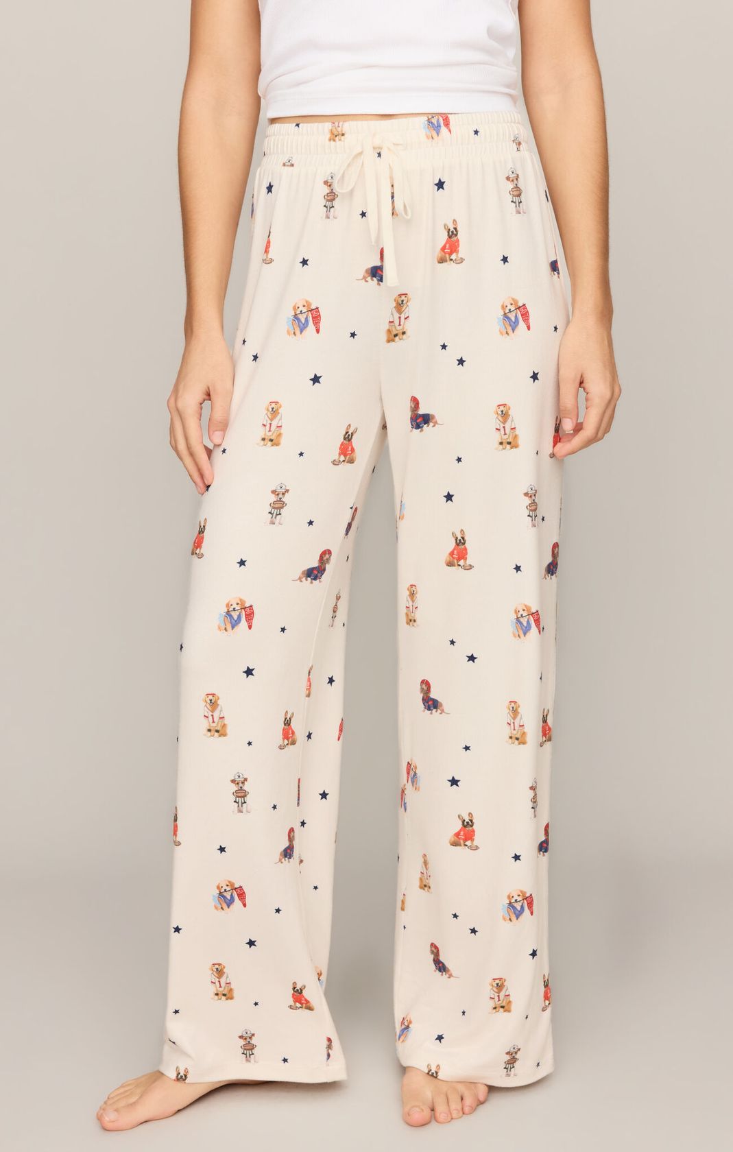 A person wearing cream-colored wide-leg pajama pants with a colorful cartoon rabbit and star print, paired with a white tank top, shown against a neutral studio background.