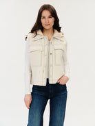 Woman wearing a cream-colored vest over a white shirt with blue jeans on a light gray background
